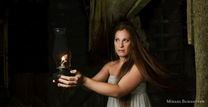 Young woman in a white dress exploring an abandoned wooden house with an oil lamp