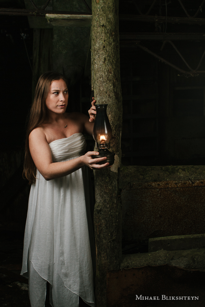 Young woman in a white dress exploring an abandoned wooden house with an oil lamp