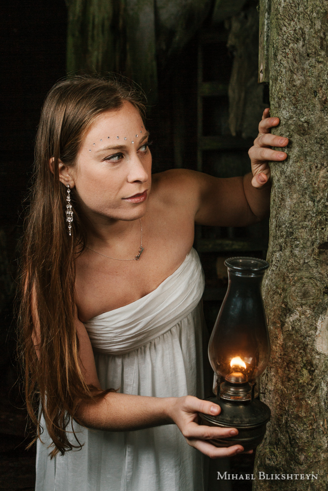 Young woman in a white dress exploring an abandoned wooden house with an oil lamp