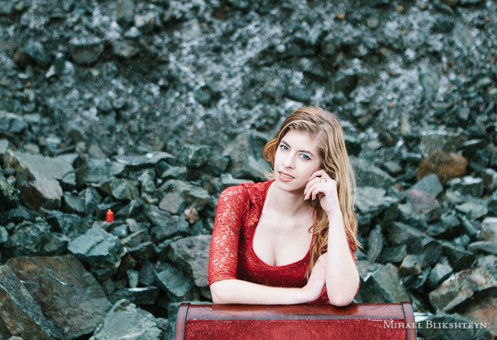 Portrait of a young woman in red dress leaning on a red velvet chair in a rock quarry with snow falling