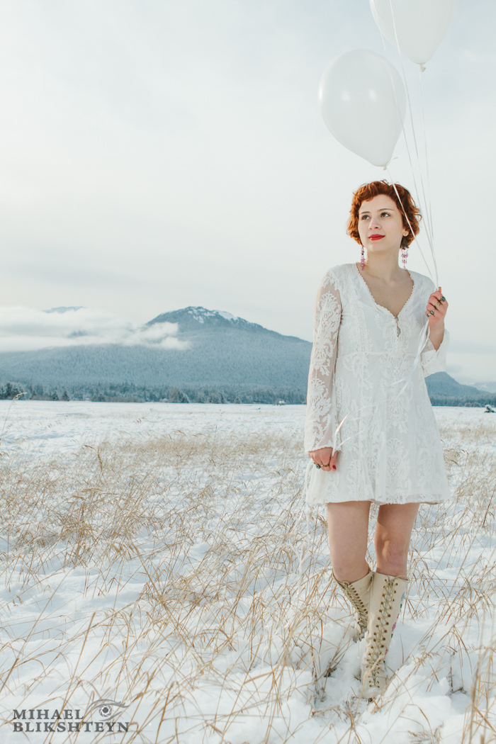 Young woman in white dress and white boots holding white balloons on a snow-covered white meadow