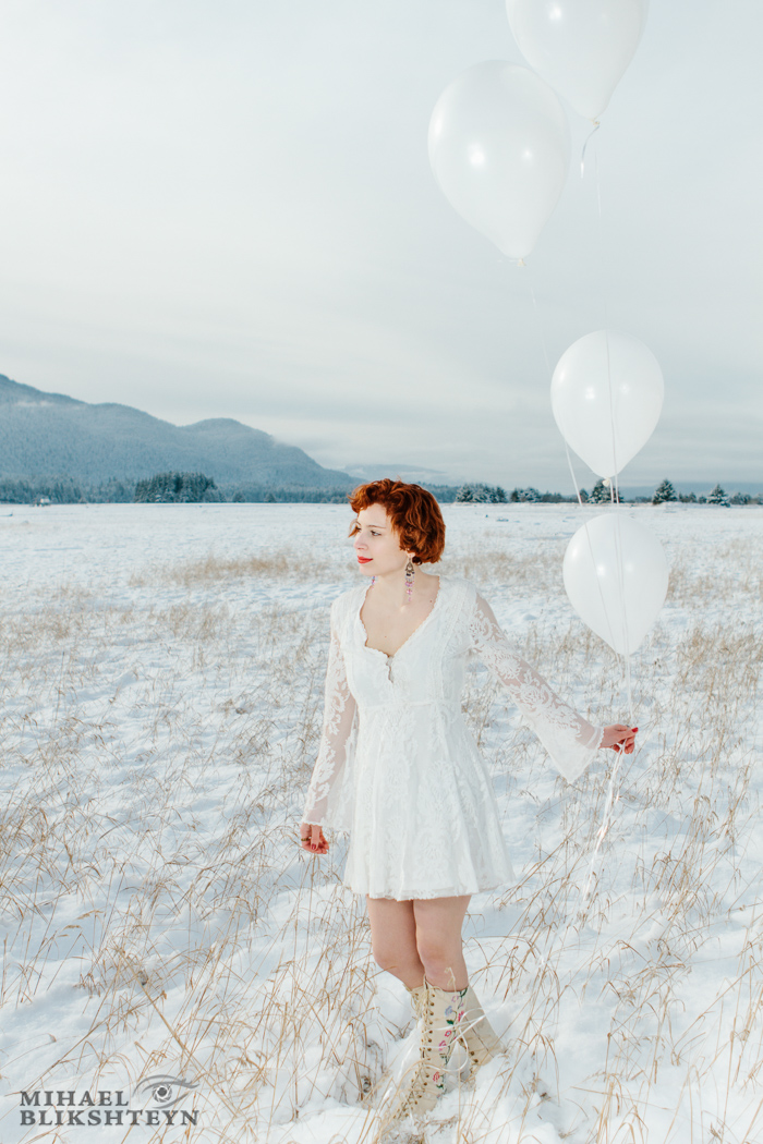 Young woman in white dress and white boots holding white balloons on a snow-covered white meadow